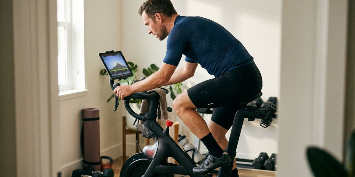 A man in athletic wear rides a black indoor stationary bike with an attached tablet screen in a bright home gym setting.