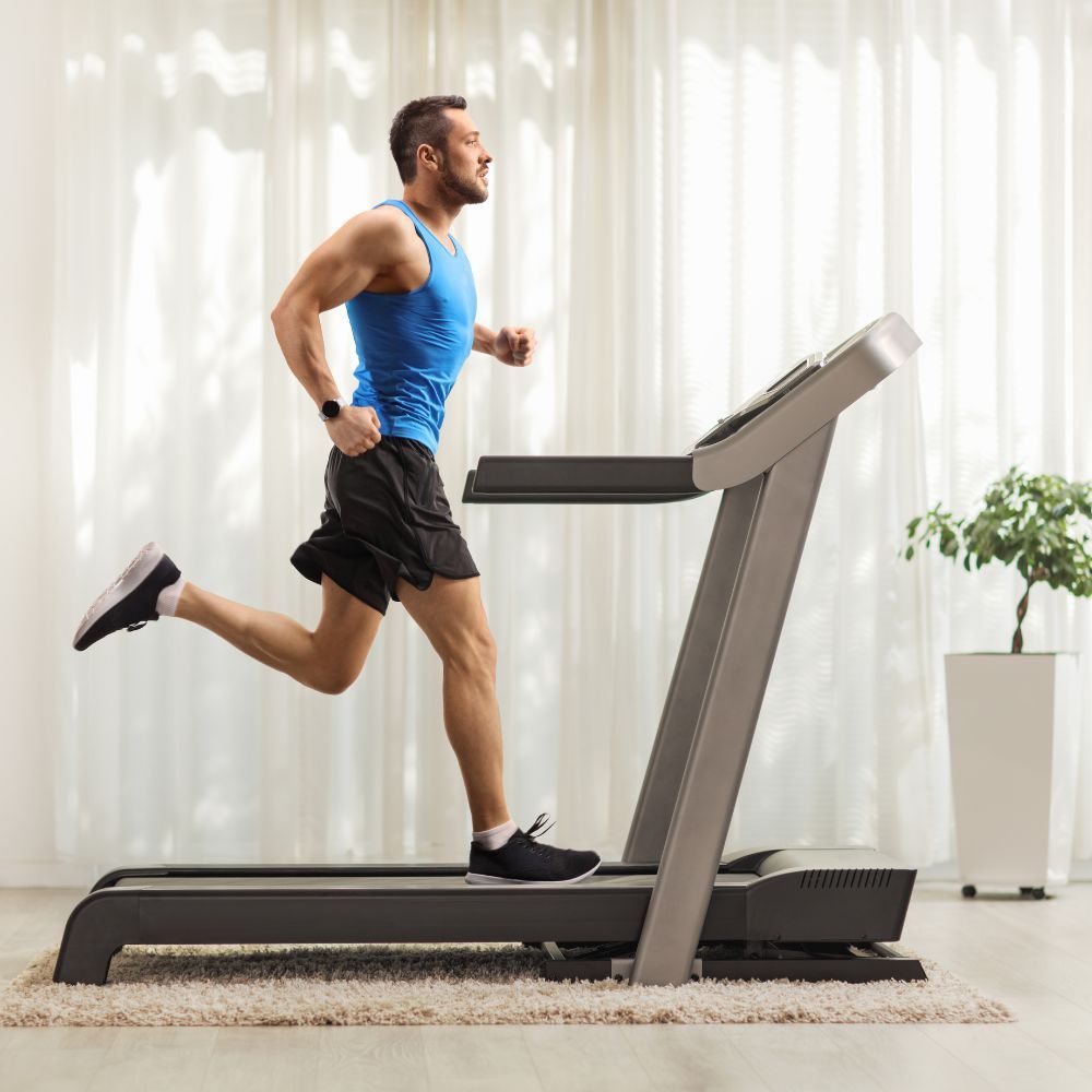 Man running on a treadmill in a home setting with curtains and a plant in the background